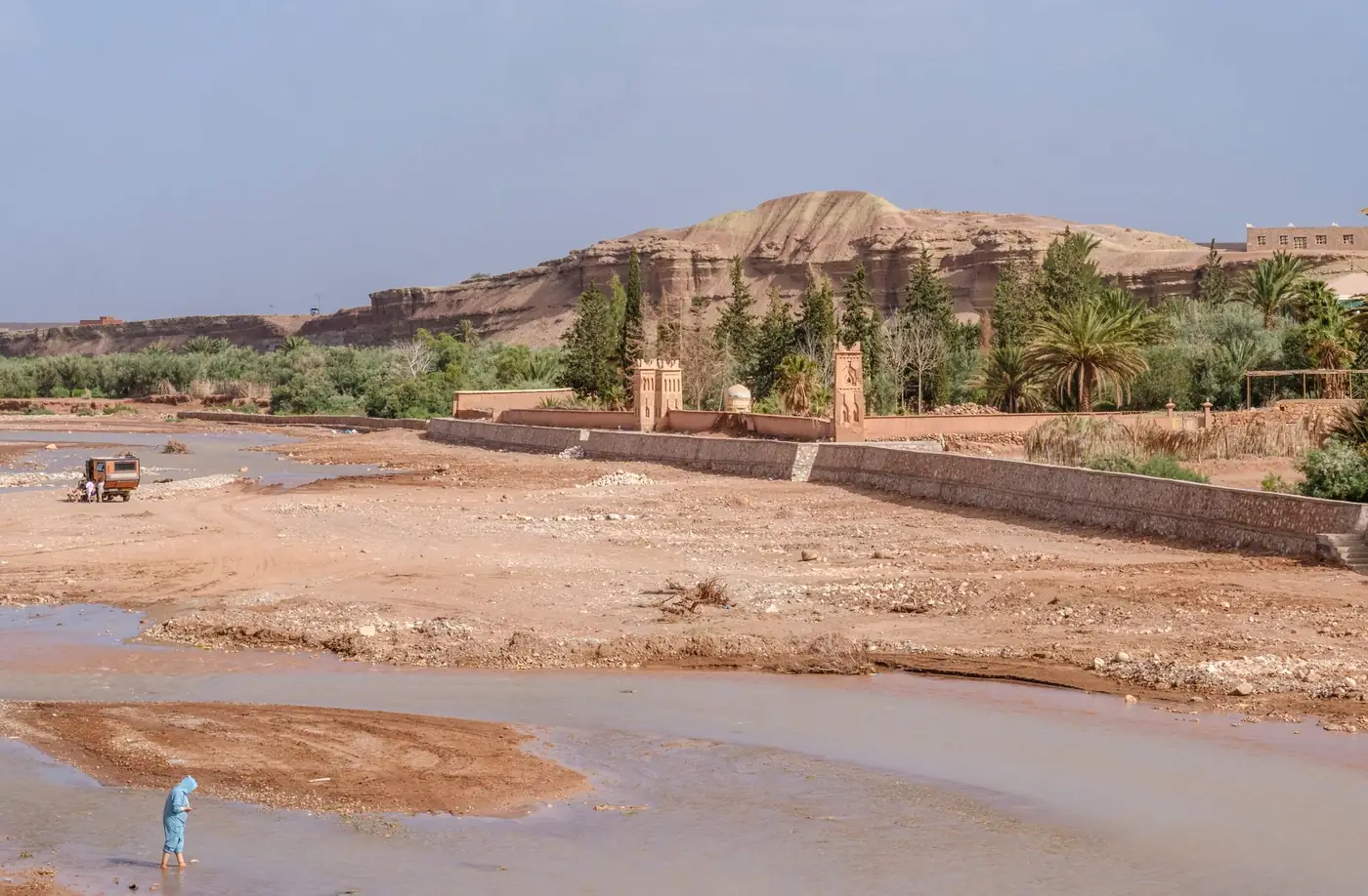 Lit de rivière au pied d'un relief ocre, avec palmiers et un mur en pisé, au Maroc.
