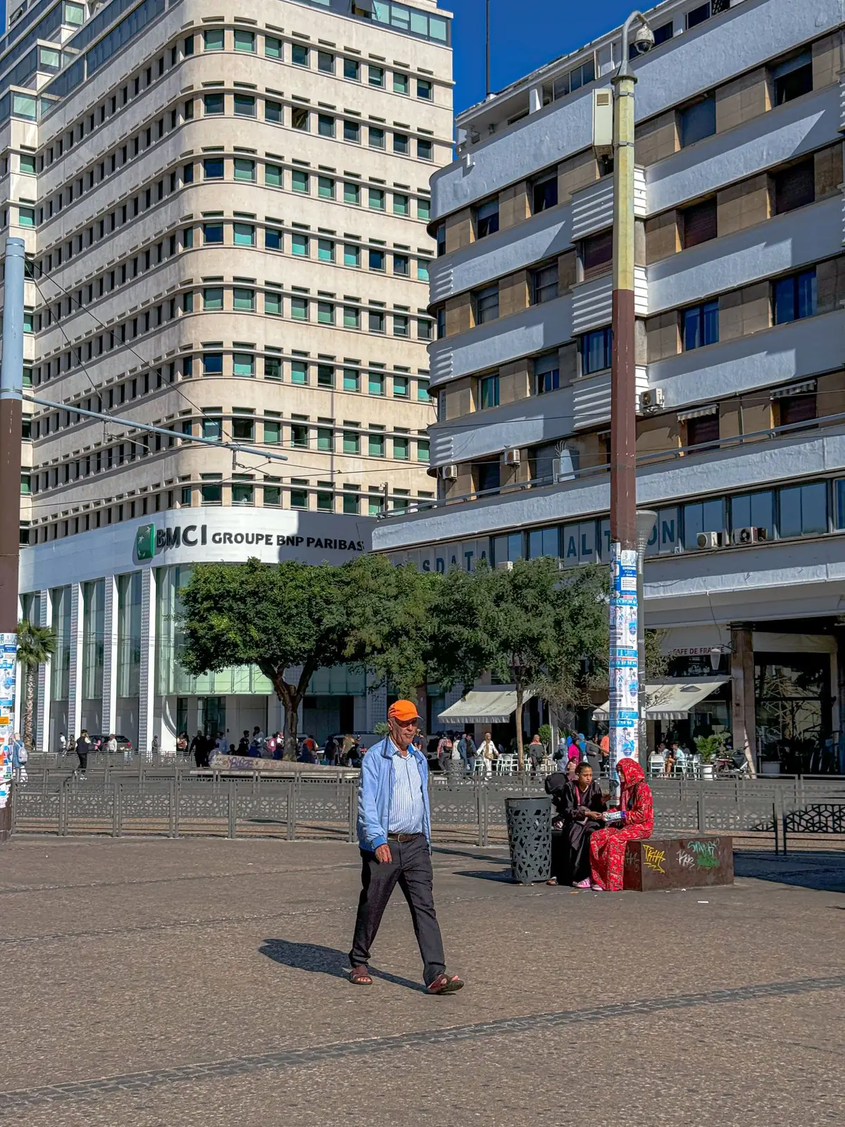Passants sur une place du centre de Casablanca, entre de hauts immeubles clairs, sous un ciel bleu.